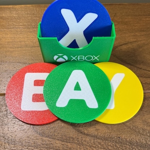 Puede incluir: Un juego de posavasos Xbox coloridos. Los posavasos son rojos, verdes, amarillos y azules con letras y logotipos blancos. También se incluye un soporte verde con el logotipo de Xbox.