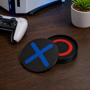 May include: A circular black coaster featuring a bright blue X symbol on its top surface, paired with a matching black circular holder that displays a vibrant red O symbol on its interior base, designed for gaming setups and drink protection.