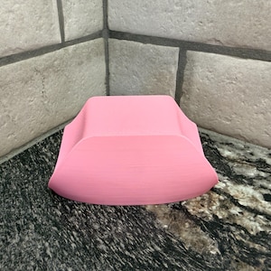 Pode incluir: Um objeto rosa sólido, curvo, com uma base arredondada e uma parte traseira elevada. O objeto é feito de um material liso e fica em uma bancada escura e salpicada. O fundo apresenta azulejos claros e texturizados.