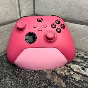 May include: A pink Xbox controller with black buttons and a black joystick. The controller has a two-tone design, with a lighter pink on the bottom half. The Xbox logo is visible at the top.