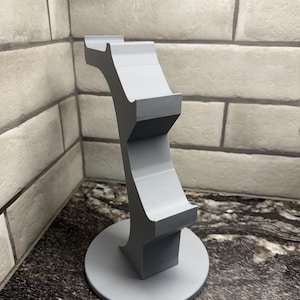 May include: A light gray, 3D-printed phone stand. The stand has a circular base, a curved central support, and two rectangular platforms. The stand is designed to hold a phone or tablet. The stand measures approximately 6 inches tall.