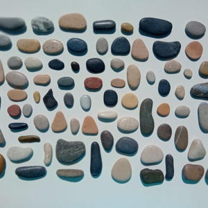 Puede incluir: Una colección de piedras lisas y redondeadas de varios tamaños y colores, incluyendo gris, beige, azul y marrón. Las piedras están dispuestas juntas, creando un patrón texturizado. La imagen muestra una estética natural y terrosa.
