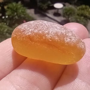 Op de afbeelding: Een gladde, amberkleurige zeeglas kei met een mat, wit bovenkant. De kei wordt in een hand gehouden, met een vage groene achtergrond.
