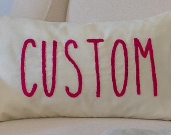 May include: Cream-coloured rectangular cushion embroidered with the word "CUSTOM" in bright pink. The cushion is on a light-coloured upholstered chair. The text is in a bold, handwritten style.