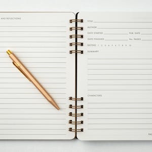 Two-page interior spread of the reading journal showing the lined “Notes and Reflections” page on the left and the structured book review layout on the right. A rose gold tone metal pen rests diagonally across the notes page. Photographed on a white background.