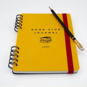 Handmade A5 yellow “Book Club Journal” with red elastic band and personalized name “Judy” on the cover, shown on an angle with a black pencil resting across the front. Wire bound with black coil and featuring a stacked books illustration printed in black ink.