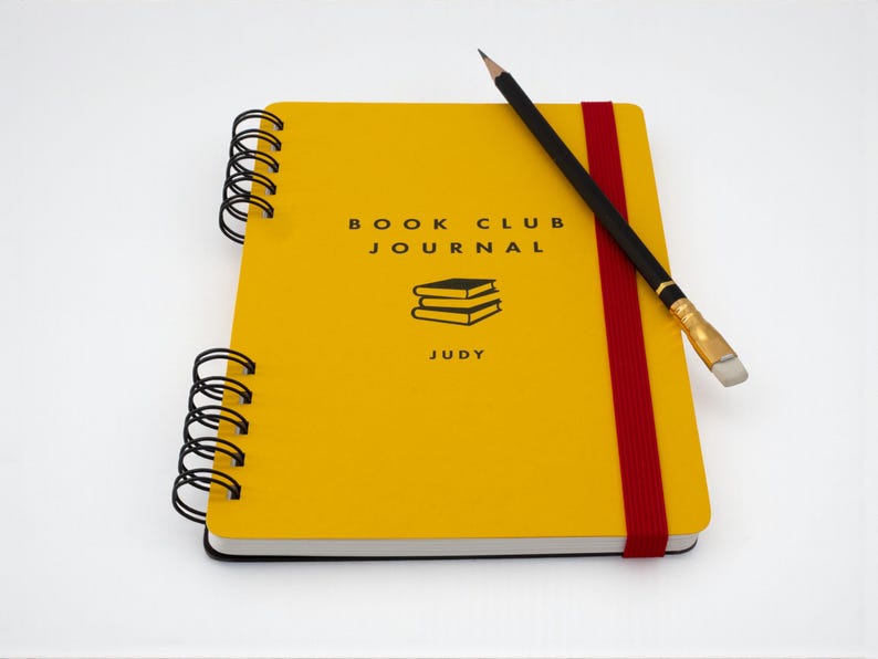 Handmade A5 yellow “Book Club Journal” with red elastic band and personalized name “Judy” on the cover, shown on an angle with a black pencil resting across the front. Wire bound with black coil and featuring a stacked books illustration printed in black ink.