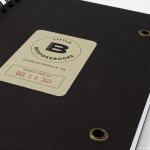 Inside back cover of the Little Bespokebooks Reading Journal showing signature “Made by hand on” label stamped in red ink with date December 15, 2025. Black cover with antique brass grommets and rounded corners. Label includes Little Bespokebooks logo and Charlottesville, Virginia mark.