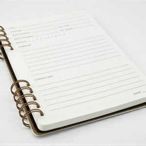 Reading journal open to a single review page with spaces for title, author, dates, rating, summary, and characters, shown at an angle against a white background.