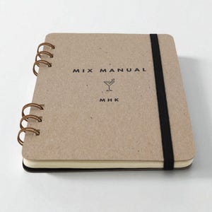 Mix Manual cocktail recipe journal with kraft chipboard cover, bronze wire binding, and black elastic band, shown closed on white background — handmade cocktail recipe notebook