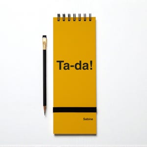 May include: A yellow spiral-bound notepad with the words "Ta-da!" in black. A black pencil with a gold ferrule rests beside the notepad. The notepad has a black horizontal stripe and the name "Sabine" at the bottom.