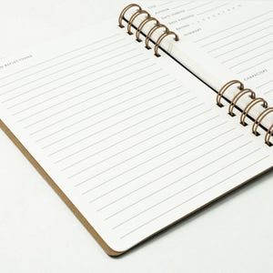 Alternate angled view of reading journal open to a full spread, showing the notes and reflections page in the foreground and the book details page in the background. Dual antique brass wire binding is visible.