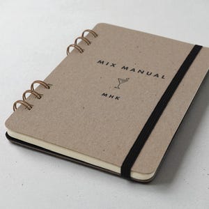 Personalized Mix Manual kraft chipboard cocktail recipe journal with black elastic band and bronze wire binding, displayed at an angle on neutral background