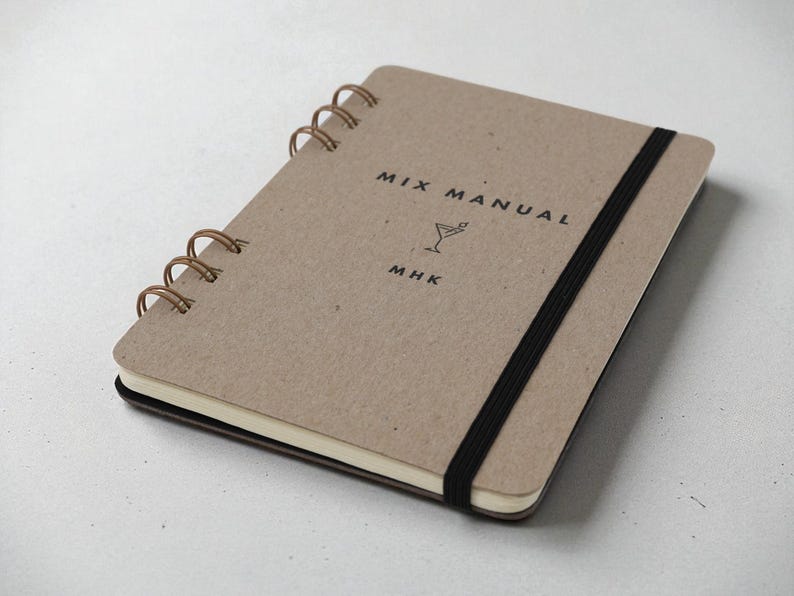 Personalized Mix Manual kraft chipboard cocktail recipe journal with black elastic band and bronze wire binding, displayed at an angle on neutral background