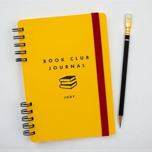 Handmade A5 yellow book club journal for groups with red elastic band and personalized name “Judy” on the cover shown with a black pencil. Wire bound with black coil and featuring a stacked books illustration printed in black ink.