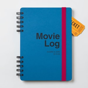 Può includere: Un taccuino a spirale blu con una chiusura elastica rosa scuro. La copertina presenta il testo "Movie Log" in grigio scuro, con "CLAIRE & JUDE" e "10.10.25" stampati sotto. Un biglietto giallo con testo nero è inserito sul lato.