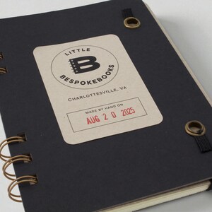 Mix Manual cocktail recipe journal with black chipboard interior cover, label stamped with date made, bronze grommets securing elastic band, and wire binding — handmade bar notebook for cocktails and mixology