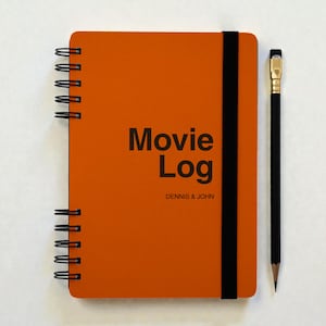 Puede incluir: Un cuaderno naranja con espiral con las palabras "Movie Log" y los nombres "DENNIS & JOHN" impresos en negro. Un lápiz negro con goma dorada está colocado junto al cuaderno.