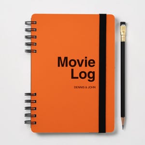 Op de afbeelding: Een oranje notitieboek met spiraalbinding en een zwarte elastische sluitband. De kaft bevat de zwarte tekst "Movie Log" en "DENNIS & JOHN". Een zwart potlood met een gouden huls en een roze gum ligt verticaal naast het notitieboek.