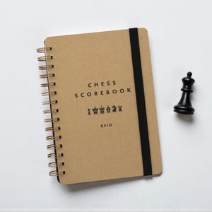 Puede incluir: Un cuaderno de anotación de ajedrez con espiral, con una cubierta de papel kraft, una banda elástica negra y el texto "CHESS SCOREBOOK" y "REID". Una reina de ajedrez negra está al lado del libro. El cuaderno mide unos 20 cm de largo.