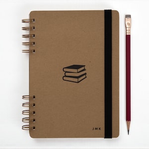 Handmade A5 book journal with kraft cover, black elastic band, and personalized initials “JMK” on the front. Spiral bound with antique brass wire and paired with a red pencil. Minimalist design featuring a small black illustration of stacked books at center.
