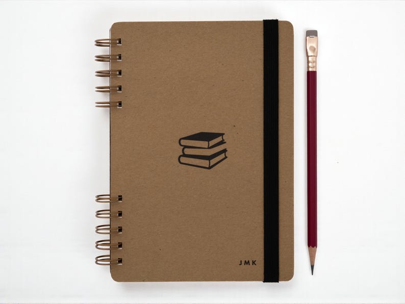 Handmade A5 book journal with kraft cover, black elastic band, and personalized initials “JMK” on the front. Spiral bound with antique brass wire and paired with a red pencil. Minimalist design featuring a small black illustration of stacked books at center.