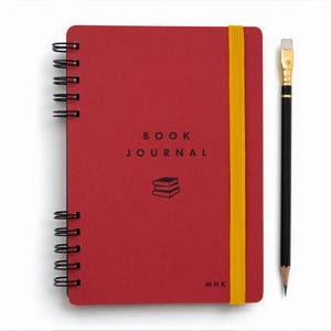 Red spiral-bound book journal with gold elastic band shown closed on a white background beside a black pencil with eraser. The cover features the title 'Book Journal' in black lettering with a small stacked-book icon below. Personal initials appear in the lower corner, emphasizing the notebook’s customizable design