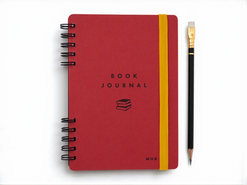 Red spiral-bound book journal with gold elastic band shown closed on a white background beside a black pencil with eraser. The cover features the title 'Book Journal' in black lettering with a small stacked-book icon below. Personal initials appear in the lower corner, emphasizing the notebook’s customizable design