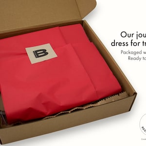 Little Bespokebooks packaging presentation. Open kraft shipping box lined with red tissue paper, sealed with a kraft sticker featuring the Little Bespokebooks “B” logo. Text in the image reads: Our journals dress for travel. Packaged with care. Ready to delight. Logo mark and Charlottesville, Virginia location also visible in the corner.