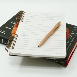 Open reading journal resting on a hardcover book, showing structured review page with a copper pen placed diagonally across the layout.