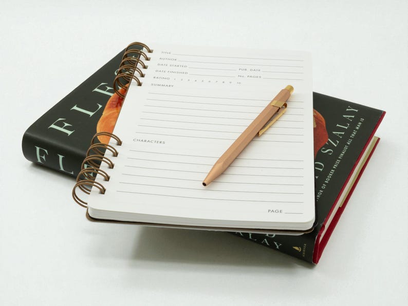 Open reading journal resting on a hardcover book, showing structured review page with a copper pen placed diagonally across the layout.