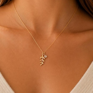 May include: A gold-toned necklace featuring a delicate chain with a pendant consisting of a small, textured leaf charm and a single, round, clear faceted stone charm suspended together from the center of the chain.