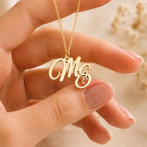 Könnte beinhalten: Eine goldfarbene Halskette mit einem Anhänger, der die in Schreibschrift verbundenen Buchstaben M und G zeigt. Der Anhänger hängt an einer zarten goldfarbenen Kette und besticht durch eine polierte metallische Oberfläche und ein fließendes Design.