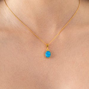 Könnte beinhalten: Goldfarbene Halskette mit einem kleinen, ovalen, blauen Anhänger. Der Anhänger hat ein helles, opalisierendes Aussehen. Die Kette ist zart und hängt am Hals einer Person. Ein schlichtes, elegantes Schmuckstück.