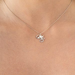 Könnte beinhalten: Eine silberfarbene Halskette mit zarter Kette. Der Anhänger zeigt die verschlungenen Schreibschriftbuchstaben "M" und "E". Die Halskette wird vor einem hautfarbenen Hintergrund präsentiert, der das Design und die Eleganz des Schmucks hervorhebt.