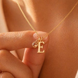 Könnte beinhalten: Eine goldfarbene Halskette mit einer zarten Kette. Die Halskette hat einen Anhänger mit dem Buchstaben "E" und einem kleinen, runden Edelstein. Die Halskette wird von einer Person gehalten.
