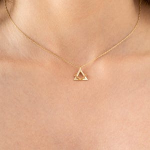 Pode incluir: Um delicado colar de ouro com um pendente geométrico. O pendente tem a forma de um triângulo com uma barra horizontal menor no interior. O colar é exibido no pescoço de uma pessoa.