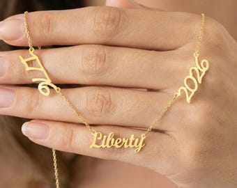 May include: A gold-colored necklace with the word "Liberty" in cursive script, flanked by the numbers "1976" and "2026". The necklace is displayed against a blurred background.