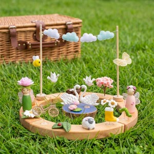 Pode incluir: Um conjunto de brinquedos de madeira caprichoso com uma base circular e figuras em miniatura. Inclui cisnes, fadas, uma abelha, uma ovelha e uma bicicleta em miniatura. A cena é montada na grama verde com uma cesta de piquenique ao fundo.