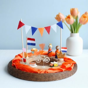 Könnte beinhalten: Eine dekorative Präsentation im niederländischen Stil mit Miniaturflaggen, einem Fahrrad und Figuren in Orange und Blau. Eine Girlande aus roten, weißen und blauen Wimpeln hängt darüber. Die Präsentation befindet sich auf einem geflochtenen Tablett mit orangefarbenem Stoff.