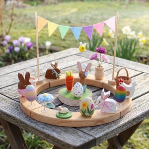 Può includere: Un'esposizione pasquale in legno con una base circolare, con uova dipinte, coniglietti, carote, fiori e una ghirlanda colorata. L'esposizione è ambientata su un tavolo di legno all'aperto.
