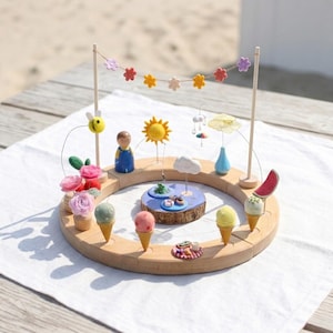 Op de afbeelding: Een ronde houten basis bevat miniatuur viltdecoraties waaronder ijshoorntjes, roze bloemen, een bij, een zon, een wolk, een blauwe vaas en een watermeloenpartje. Een slinger van kleurrijke viltbloemen hangt tussen twee houten stokjes boven het geheel.