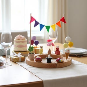 Pode incluir: Uma mesa festiva com um bolo, presentes e uma peça central de madeira decorada com artigos de festa em miniatura. Uma faixa de arco-íris e balões contribuem para o ambiente festivo. Taças de vinho e pratos também estão presentes.