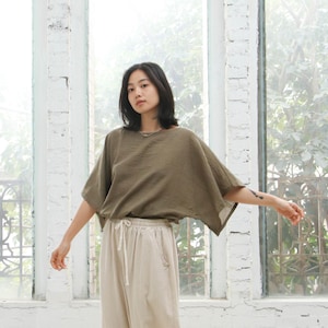 Peut inclure: Un haut vert olive à manches courtes et amples, de coupe ample. Il est associé à un pantalon beige clair à taille élastique. L'ensemble est présenté devant un mur de briques blanches et une grande fenêtre.