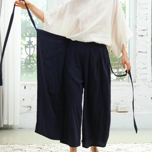 May include: A woman wearing a white top and dark blue wide-leg trousers with a wrap-around tie. The trousers have a relaxed fit and are made of a lightweight fabric.