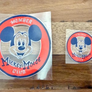Puede incluir: Dos pegatinas de miembro del Mickey Mouse Club. Las pegatinas presentan la cara de Mickey Mouse en el centro, rodeada por un círculo rojo con las palabras "MEMBER" y "Mickey Mouse CLUB" en azul. Una pegatina es más grande que la otra.