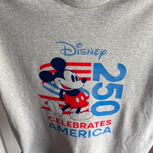 May include: A heather grey t-shirt featuring a graphic of Mickey Mouse in red shorts. The design includes the text Disney 250 and CELEBRATES AMERICA in blue and red, with stylised red and blue stripes behind the character on the front of the shirt.