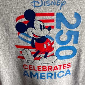 May include: A heather grey t-shirt featuring a graphic of Mickey Mouse in front of red and blue horizontal stripes. The text reads Disney at the top, 250 in large blue font on the right, and CELEBRATES AMERICA in red and blue at the bottom.