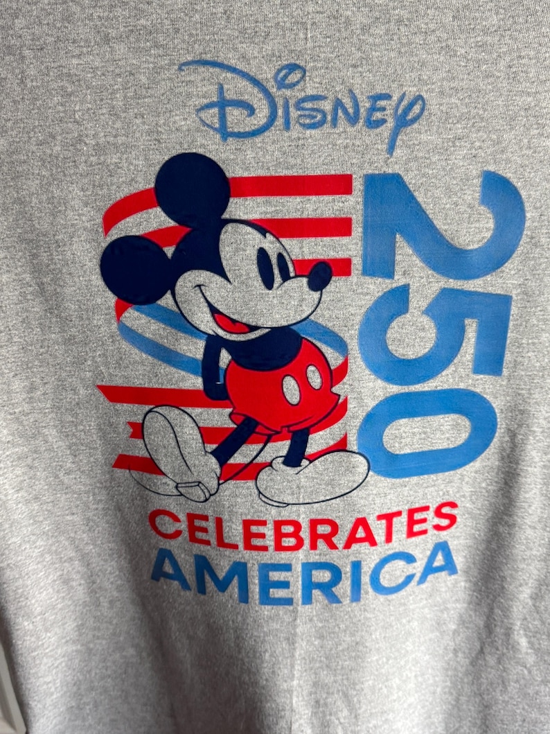 May include: A heather grey t-shirt featuring a graphic of Mickey Mouse in front of red and blue horizontal stripes. The text reads Disney at the top, 250 in large blue font on the right, and CELEBRATES AMERICA in red and blue at the bottom.