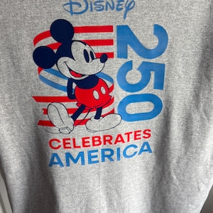 May include: A heather grey t-shirt featuring a graphic of Mickey Mouse in red shorts. The design includes the blue text Disney, the number 250 in large blue font, and the red and blue text CELEBRATES AMERICA over red and blue wavy stripes.